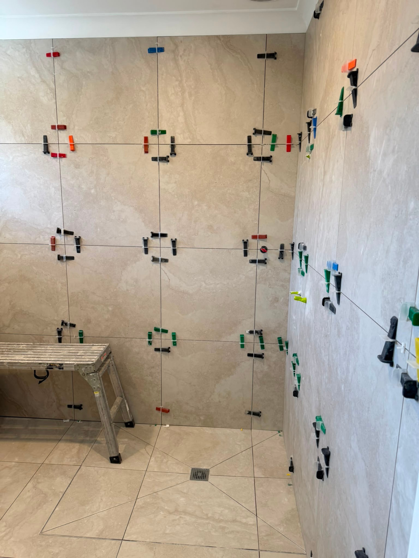 Travertine-look wall tiles held in levelling clips during installation above a tiled shower floor